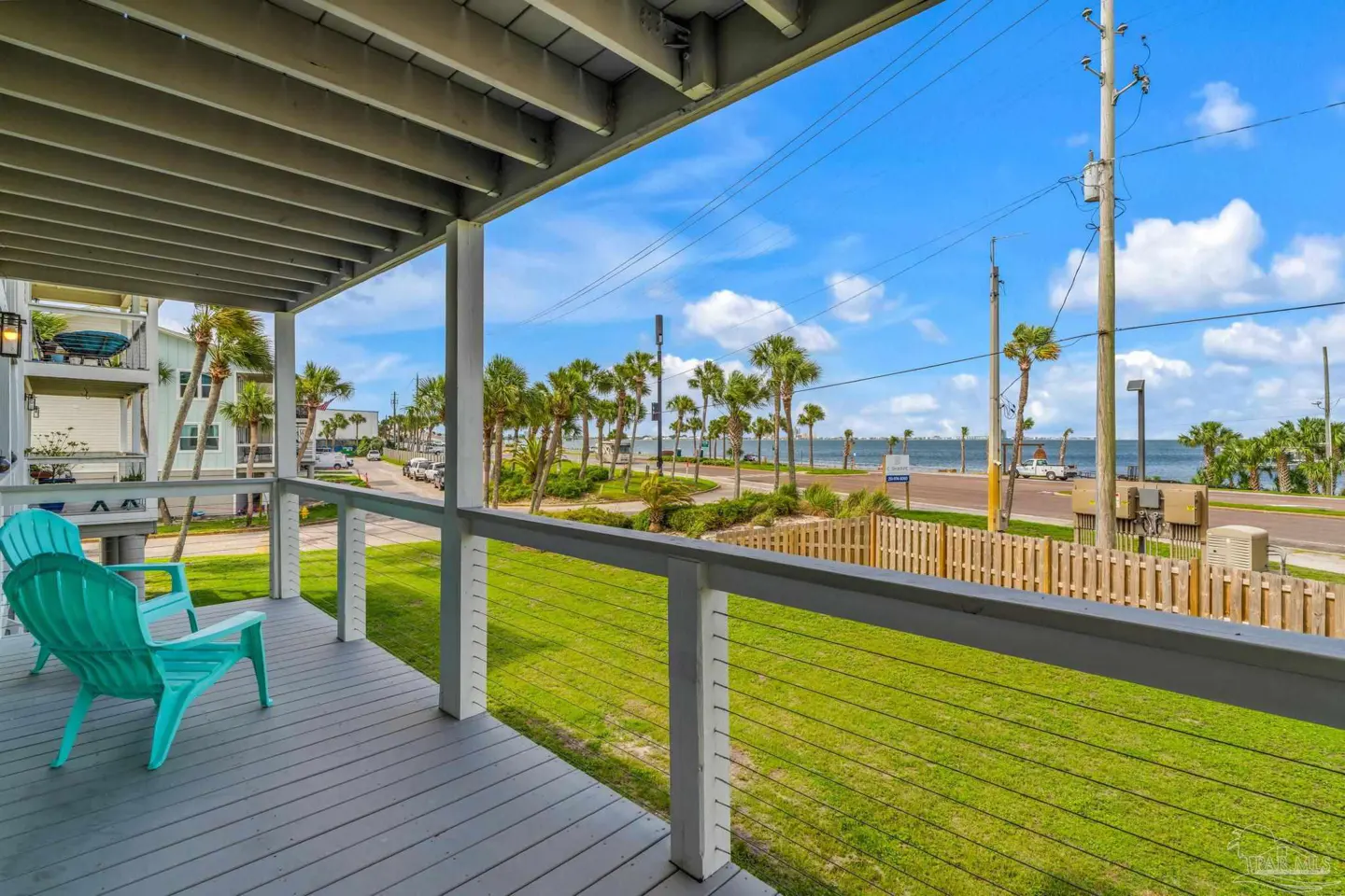 View from a covered wooden deck with two turquoise chairs overlooking a grassy yard, palm trees, and a distant bay.
