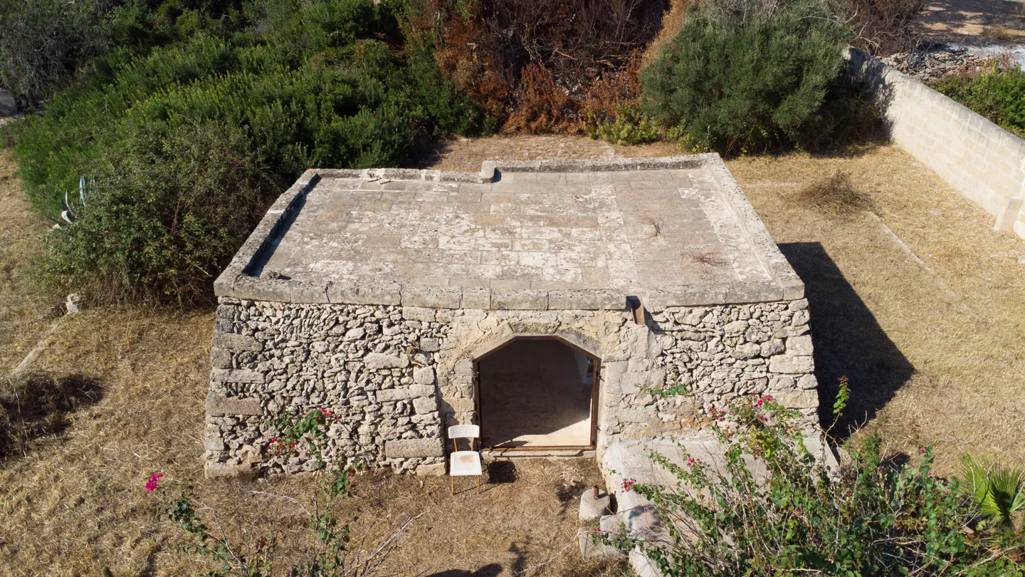 Aerial view of a stone building with an arched doorway and a white chair in front, surrounded by greenery.