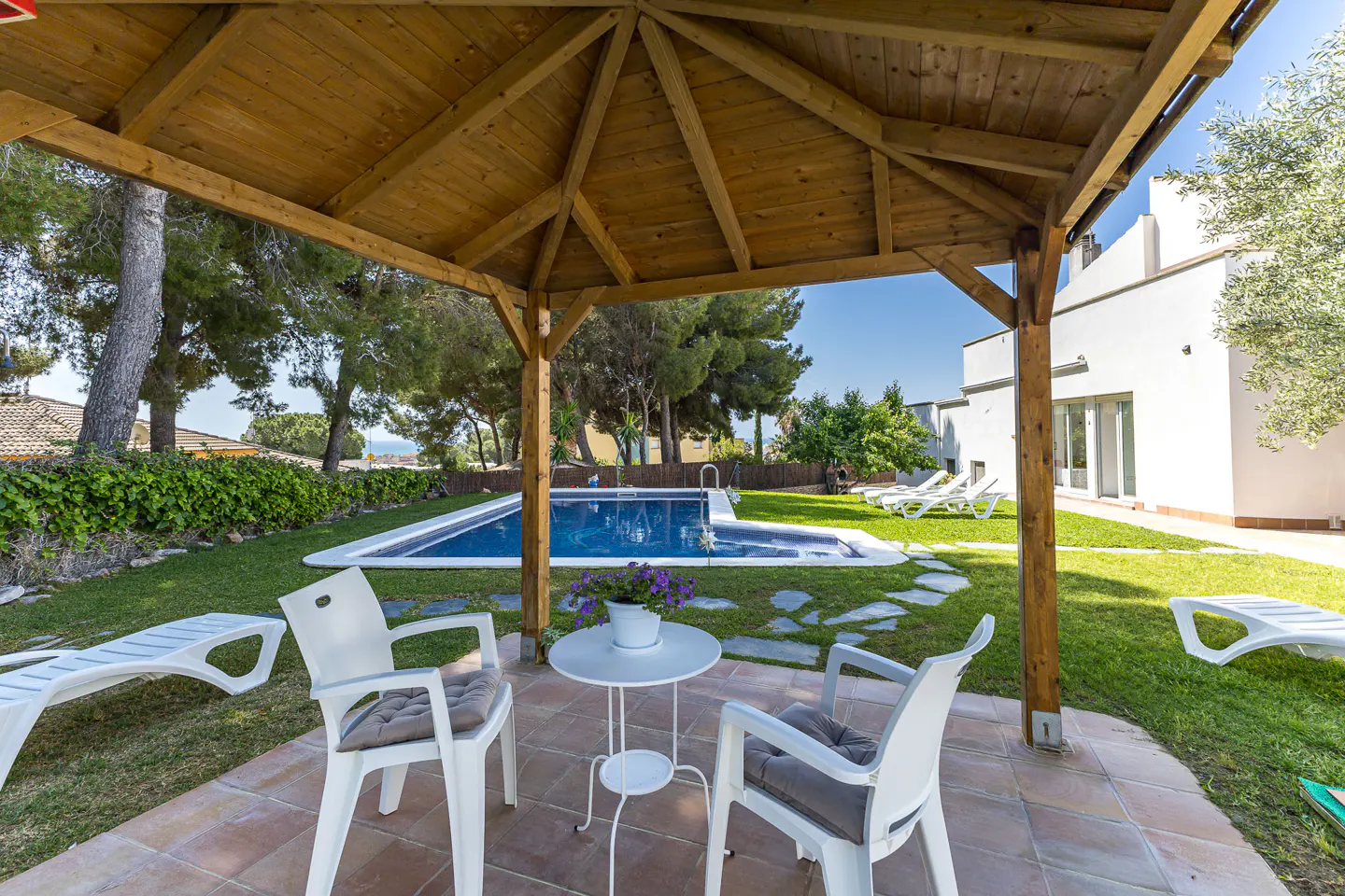 Outdoor patio with a wooden gazebo, two white chairs, a small table with flowers, and a blue swimming pool in the background.