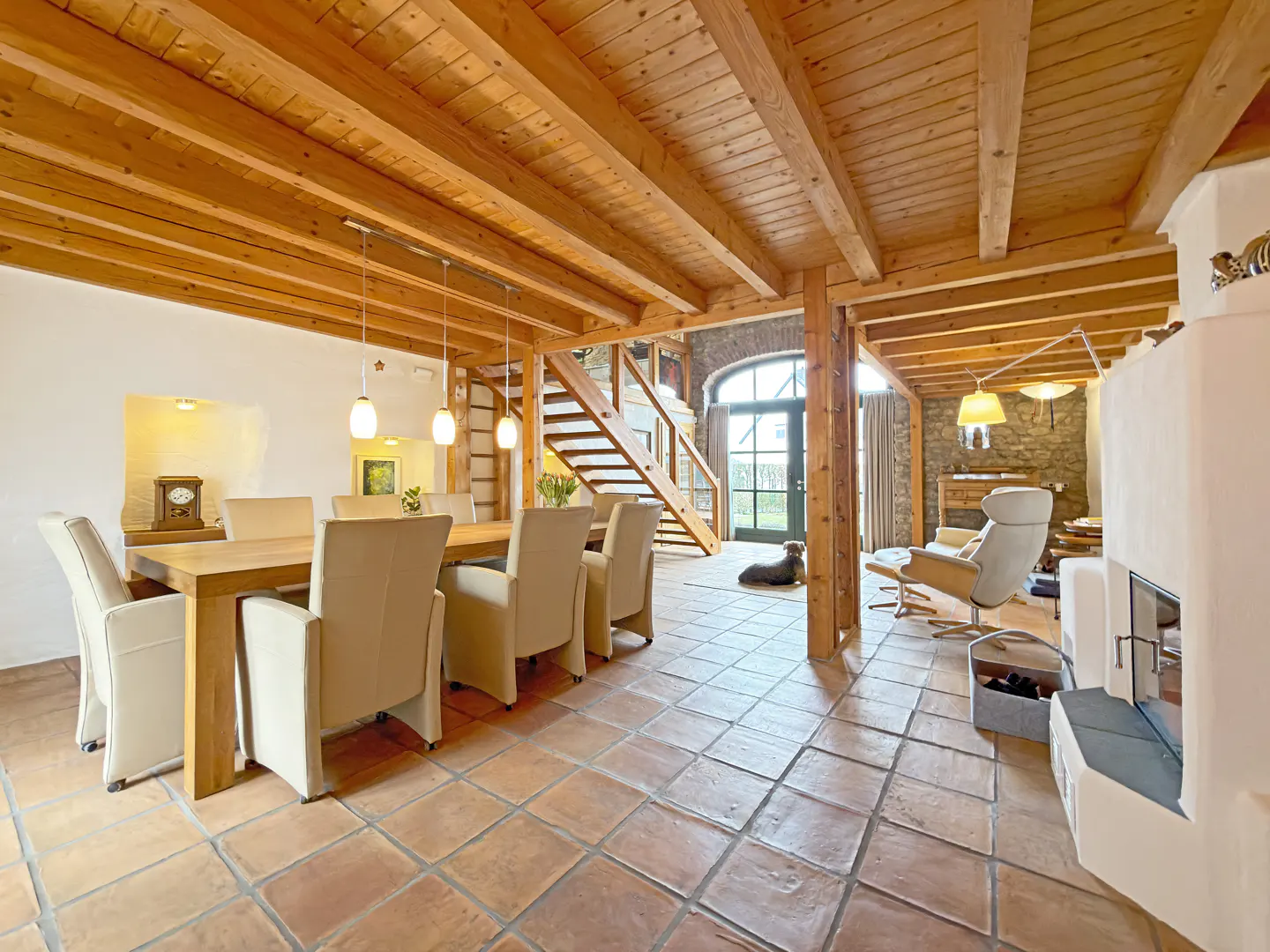 Open-concept living space with wood beams, tile floors, dining table, and fireplace. A staircase leads to the upper level.