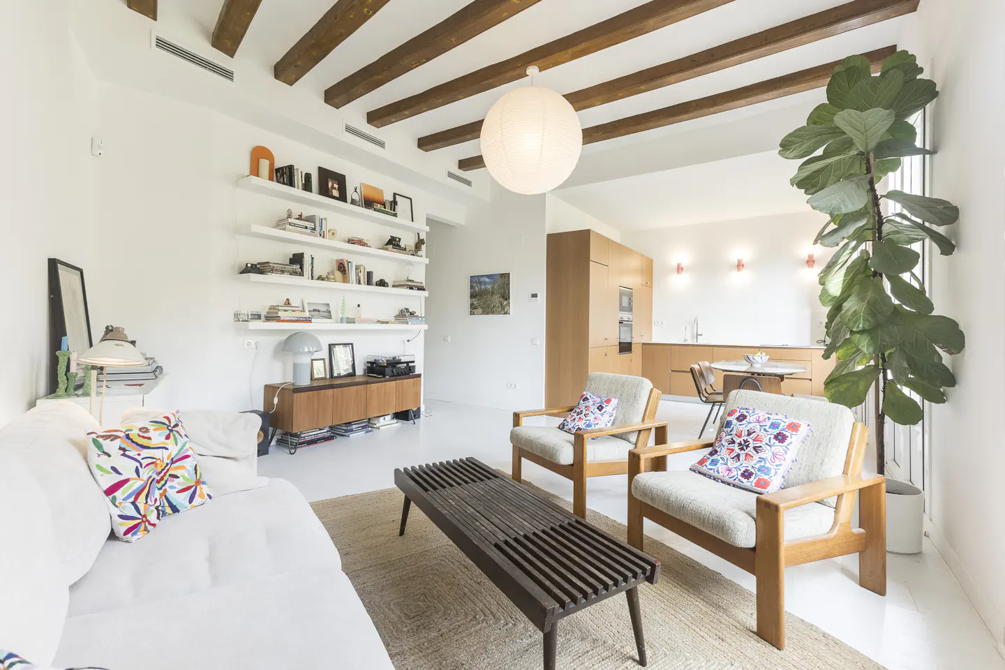 Bright living room with white walls, wooden beams, and a large round light fixture. A white sofa, wooden chairs, and a bench sit on a jute rug. Shelves line the wall.