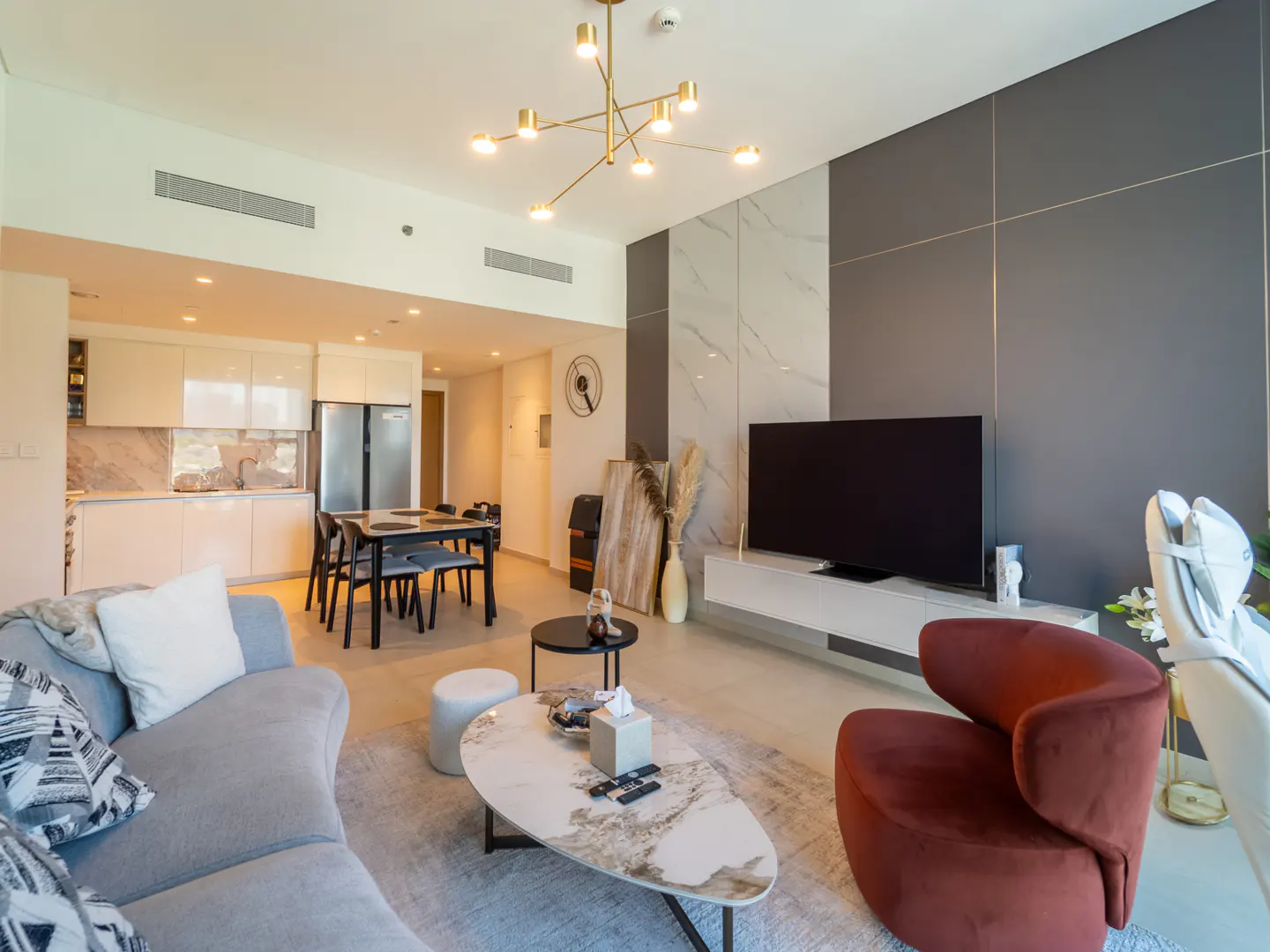 A modern apartment interior with a gray sofa, brown chair, and open kitchen. A gold chandelier hangs above.