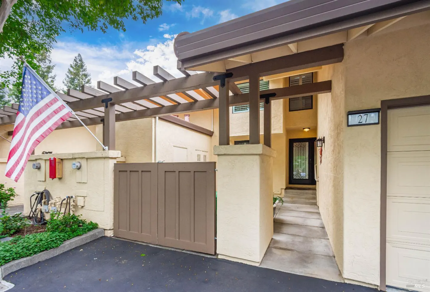 Exterior view of a tan house with a brown pergola, gate, and trim. An American flag is displayed on the left. The house number "27" is visible.