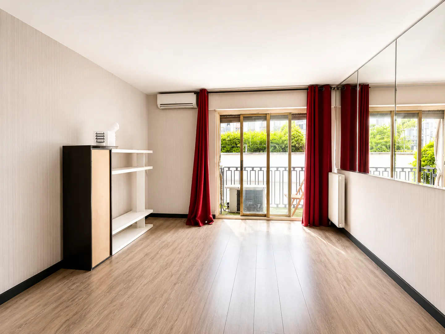 Empty room with light wood floors, white walls, a black shelving unit, red curtains, and a mirrored wall facing a balcony.