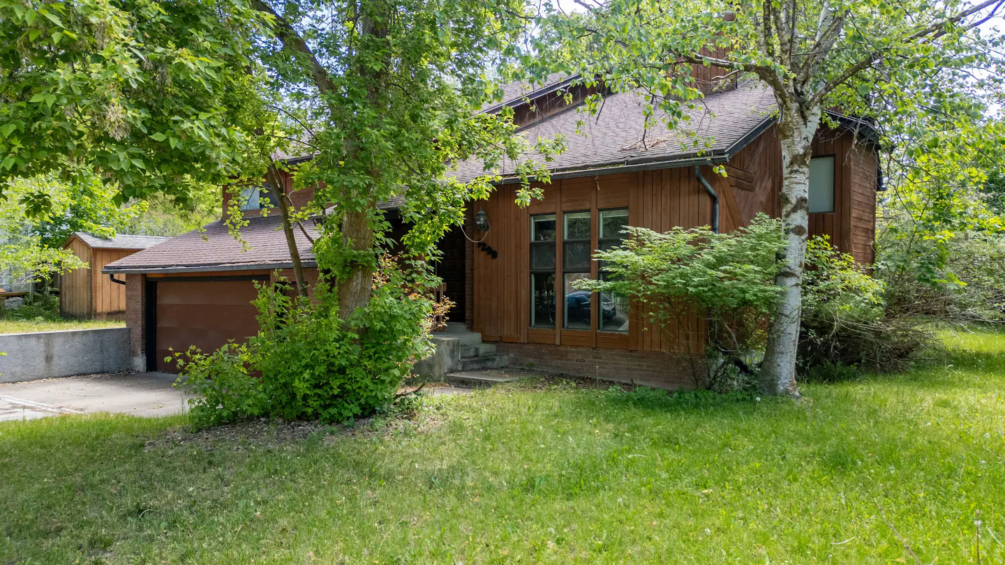 A brown, two-story house with a garage is surrounded by green trees and grass.