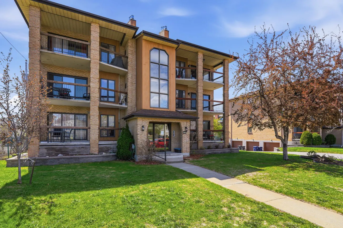 Three-story brick apartment building with balconies, a green lawn, and a sidewalk leading to the entrance.