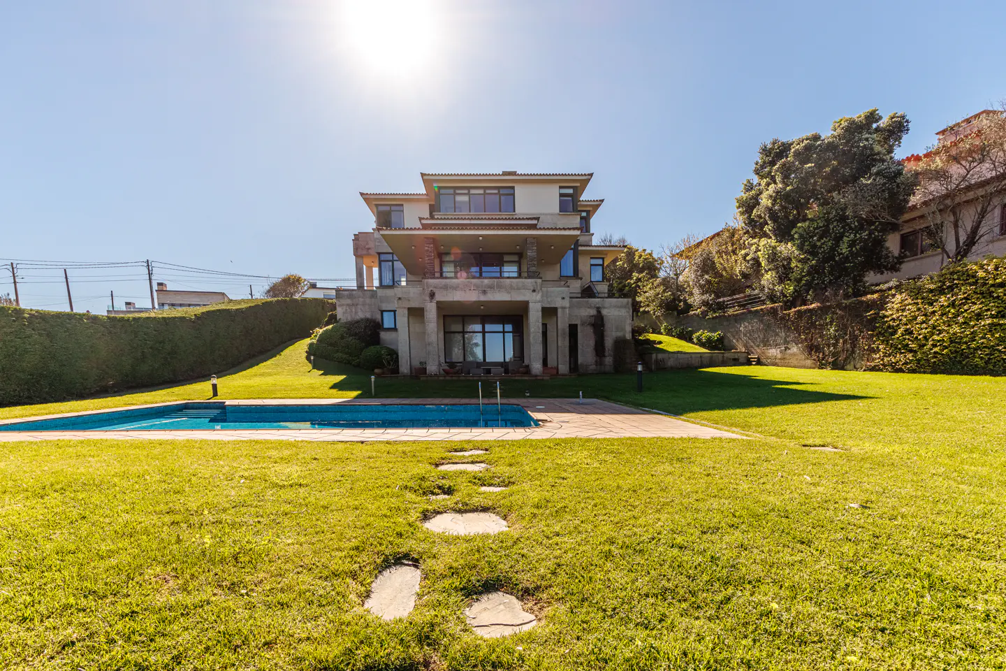A modern, multi-story house with a pool and green lawn on a sunny day. Stone path leads to the pool.