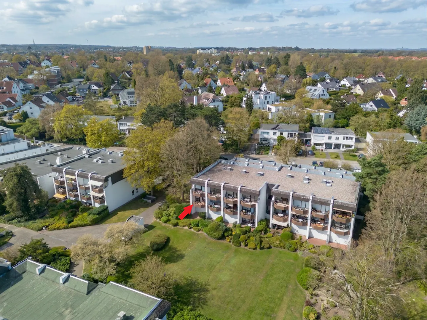 Aerial view of a white apartment building with balconies, surrounded by green lawns and trees. A red arrow points to a specific balcony.