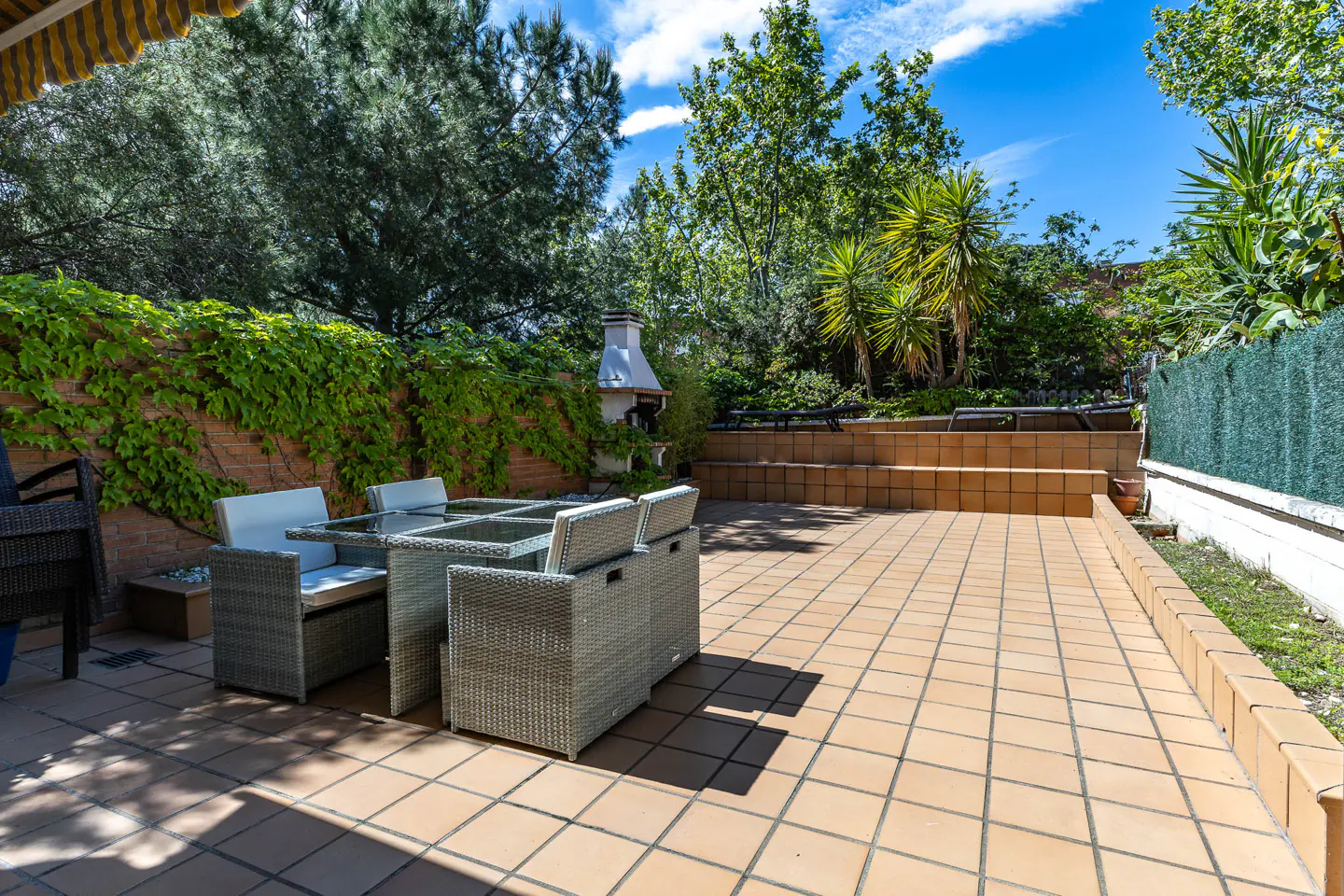Outdoor patio with wicker table and chairs on a tiled floor, surrounded by lush greenery and a brick wall with ivy.