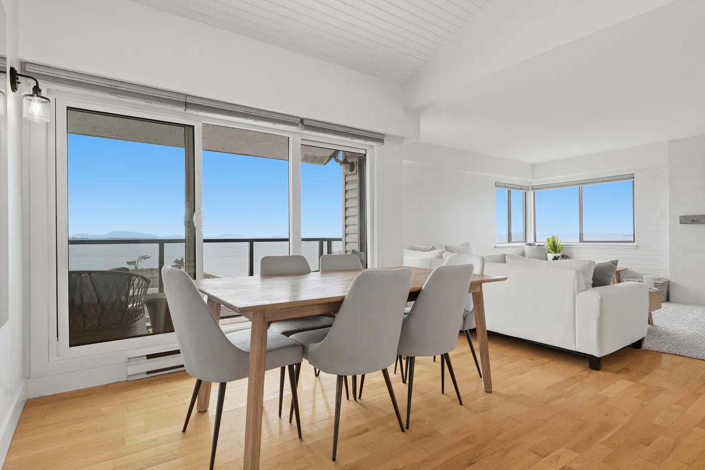 Bright, modern dining area with a wood table, gray chairs, and a sliding glass door to a balcony with an ocean view. White walls and wood floors.