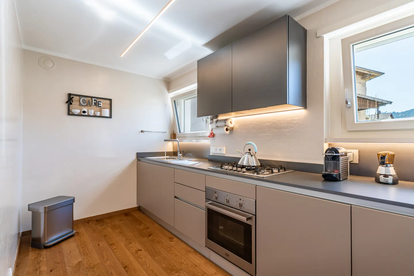 Modern kitchen with gray cabinets, stainless steel appliances, and wood floors. A kettle sits on the stovetop. Natural light streams through the window.