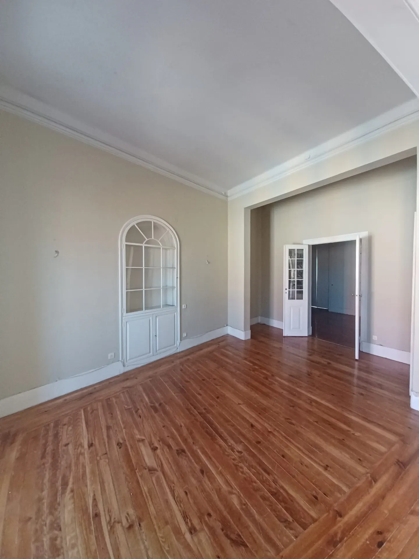 Empty room with hardwood floors, light beige walls, and white trim. A white arched cabinet is on the left wall, and an open doorway is on the right.
