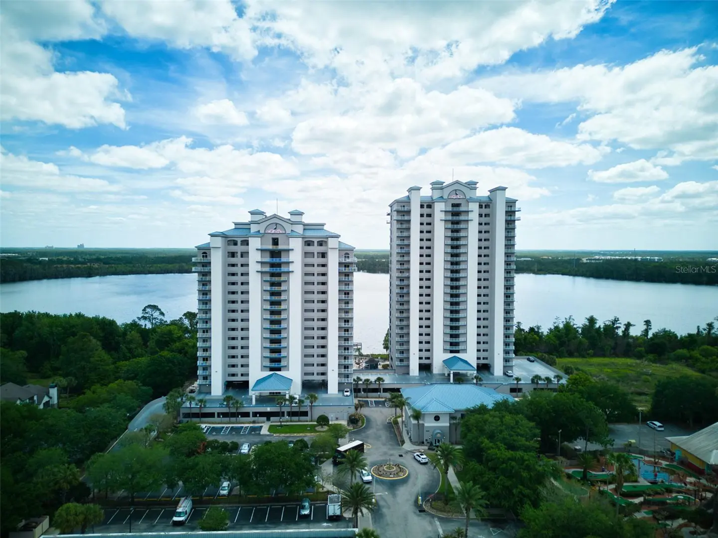 Aerial view of two tall, white condo buildings with blue accents, surrounded by green trees and a lake under a cloudy sky.