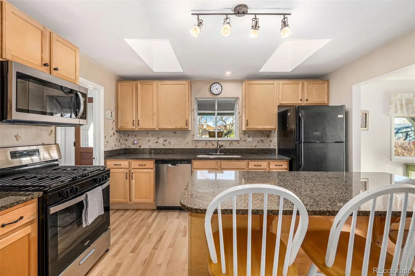 Bright kitchen with light wood cabinets, granite countertops, stainless steel appliances, and a center island with white chairs.