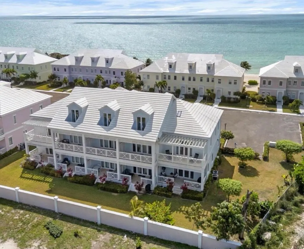 Aerial view of pastel-colored townhouses with white roofs, balconies, and a white fence, near a turquoise ocean.