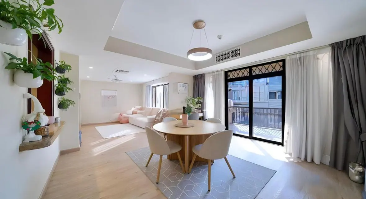 Bright, modern living space with a round dining table, four chairs, and a light gray rug. A white sofa is visible in the background. Plants adorn the wall.