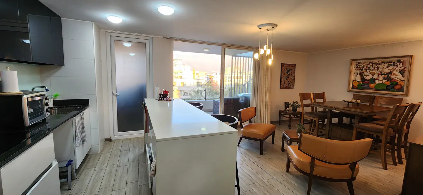 A bright apartment interior with a kitchen, dining area, and balcony. The kitchen has black countertops and white cabinets. The dining area has a wooden table and chairs.