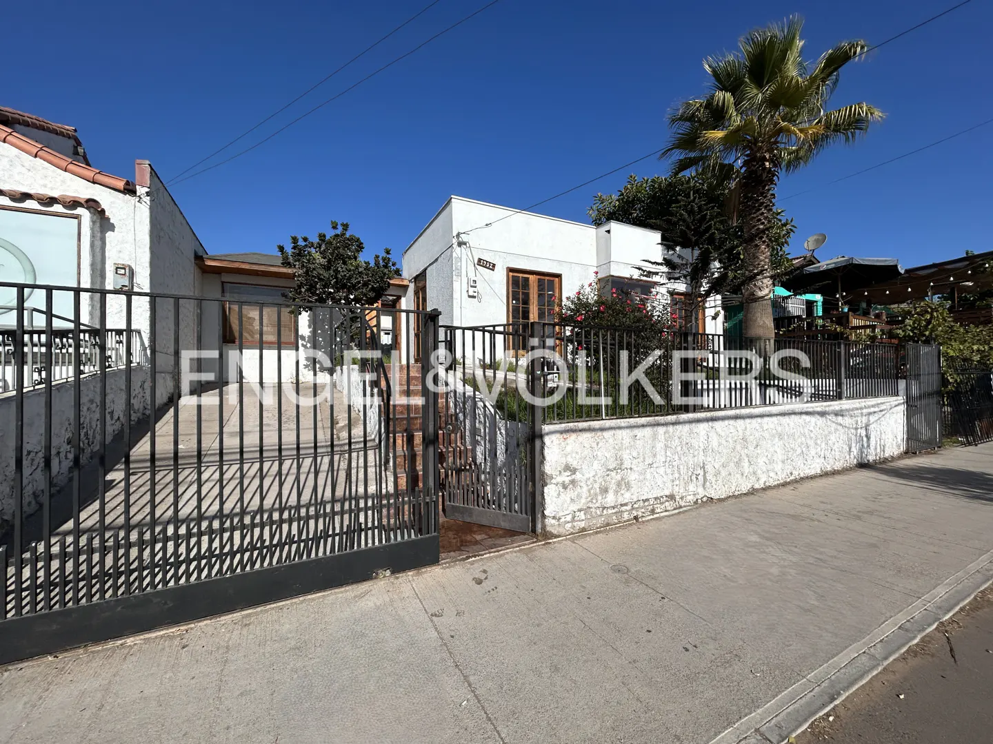 Exterior view of a white stucco house with a black metal fence and a palm tree against a blue sky.