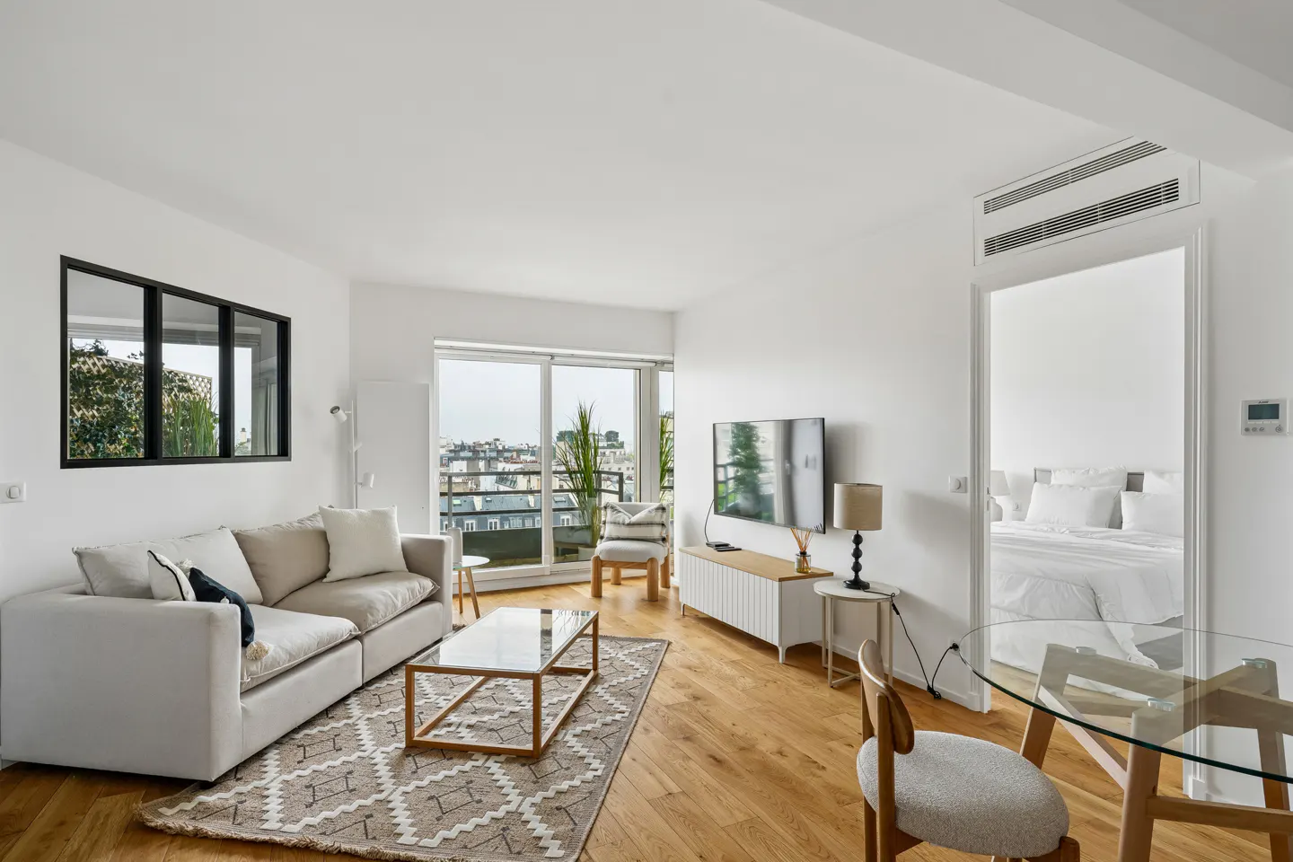 Bright, modern living room with a beige sofa, glass coffee table, and wood floors. A bedroom is visible through an open doorway.
