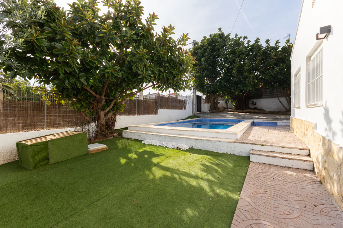 Backyard with a blue swimming pool, green lawn, trees, and a white house with a stone walkway.