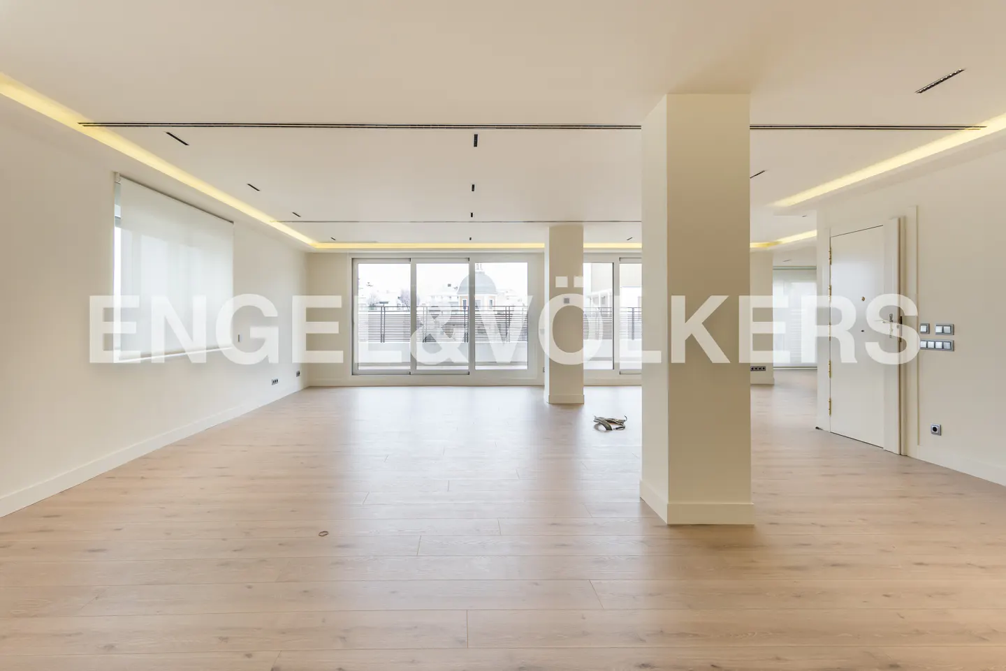 Bright, empty room with light wood floors, white walls, and large windows leading to a balcony. Column in the center.
