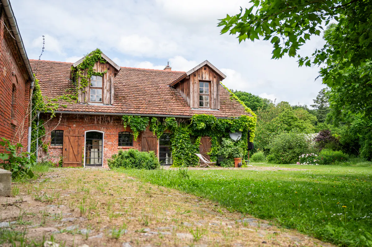 A two-story brick house with a red tile roof is partially covered in green vines. The house has wooden shutters and dormer windows, and is surrounded by a green lawn.
