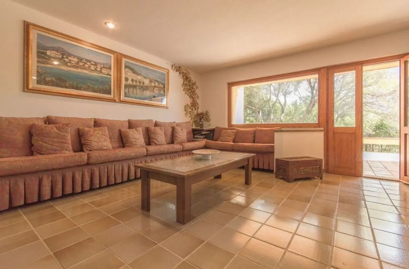 Living room with terracotta tile floor, long brown sofa with pillows, wood coffee table, and paintings on the wall. Doors open to the outside.
