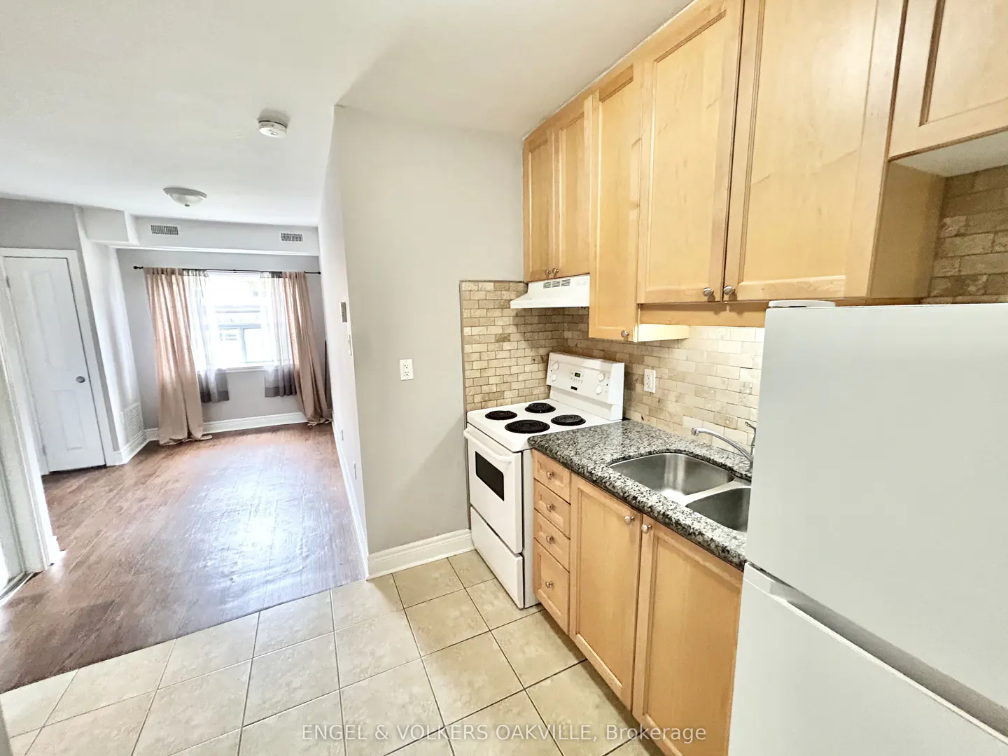 A bright kitchen with light wood cabinets, granite countertops, and a white stove. A living room is visible in the background.