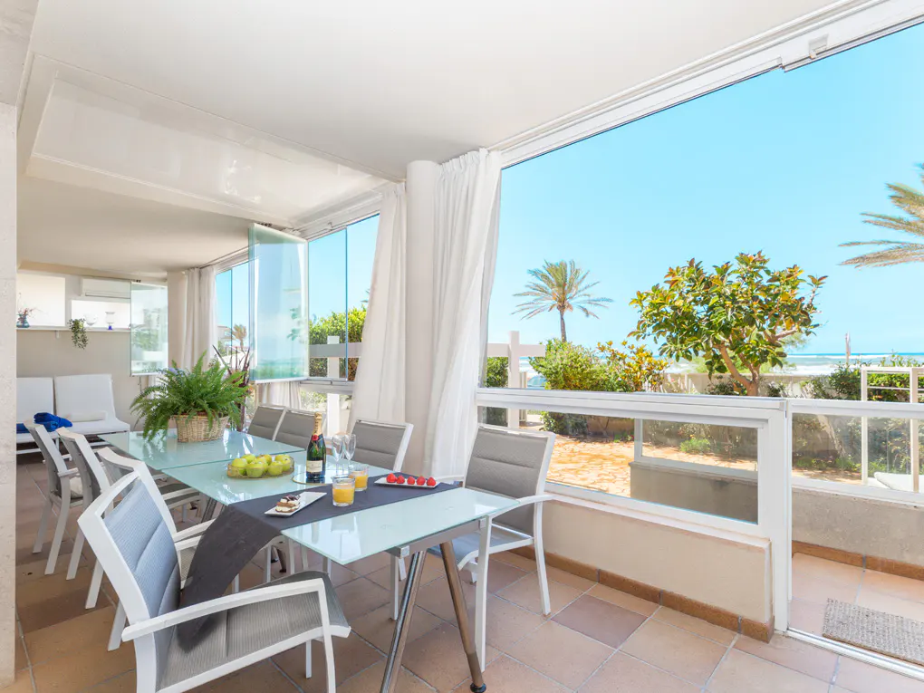 Bright patio with a glass table, gray chairs, and a view of the beach with palm trees.