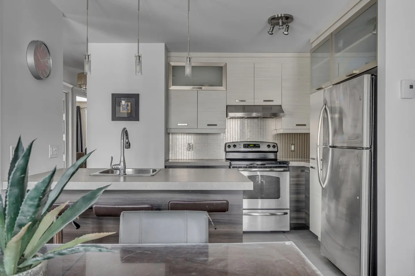 A modern kitchen with stainless steel appliances, light gray cabinets, and a breakfast bar with stools. A potted plant sits on a table in the foreground.