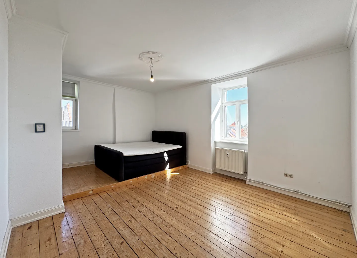 Bright, empty bedroom with wood floors, white walls, and a black bed. A window lets in natural light.