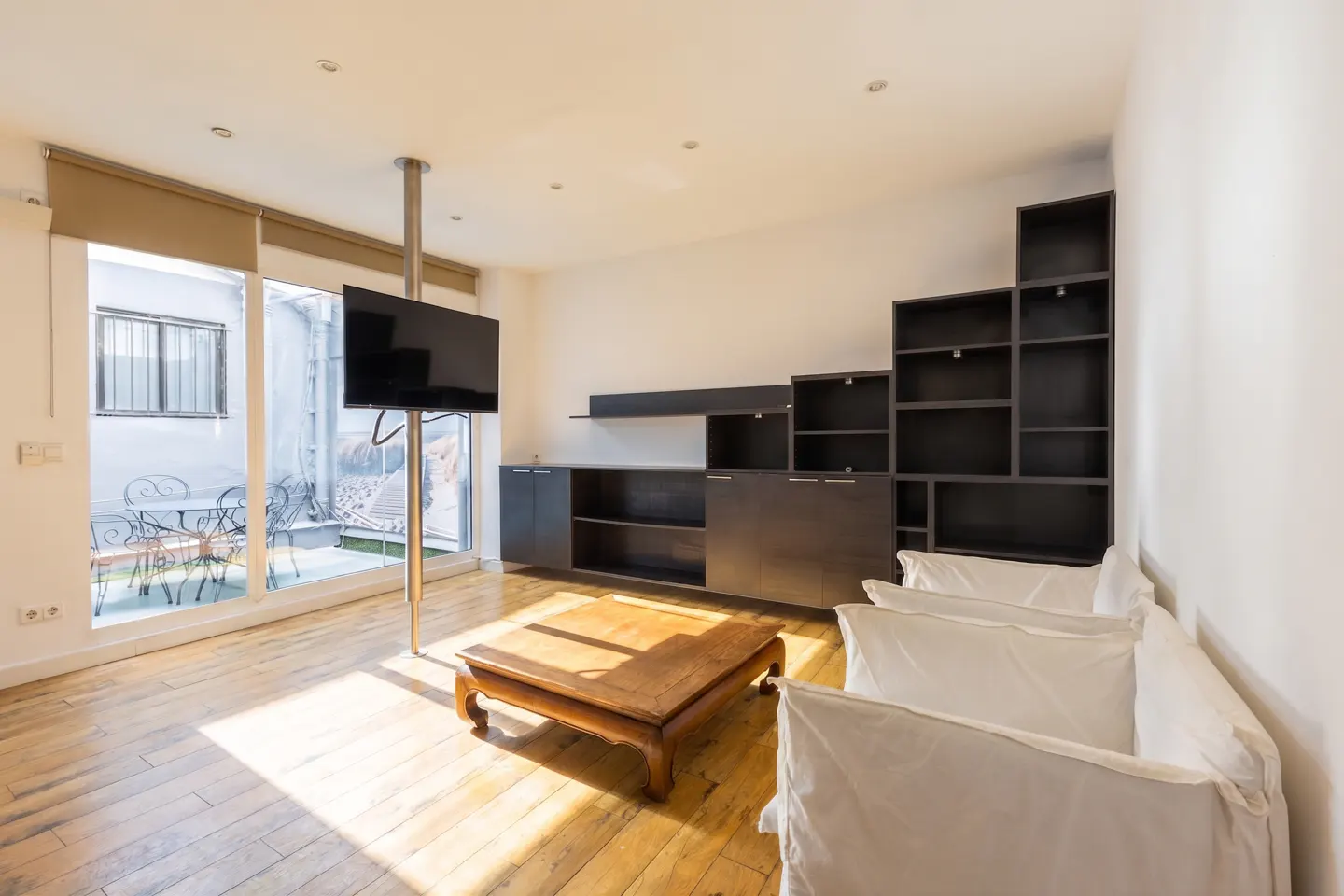 Bright living room with wood floors, white sofa, and a wooden coffee table. A TV is mounted on a pole. A patio is visible through a glass door.