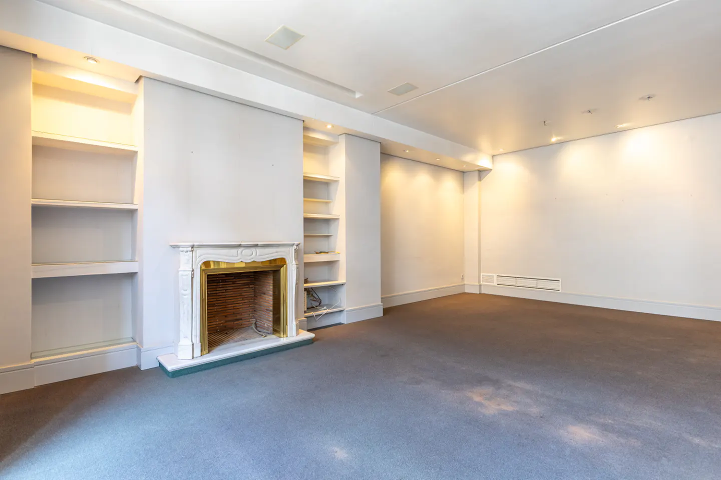 Empty room with gray carpet, white walls, and a fireplace with gold trim flanked by built-in shelves.