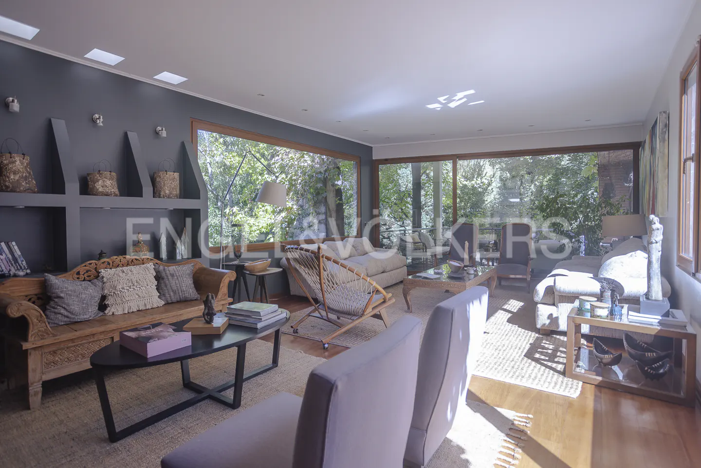 Bright living room with large windows overlooking greenery. Gray walls, wood floors, and neutral furniture create a cozy, modern space.