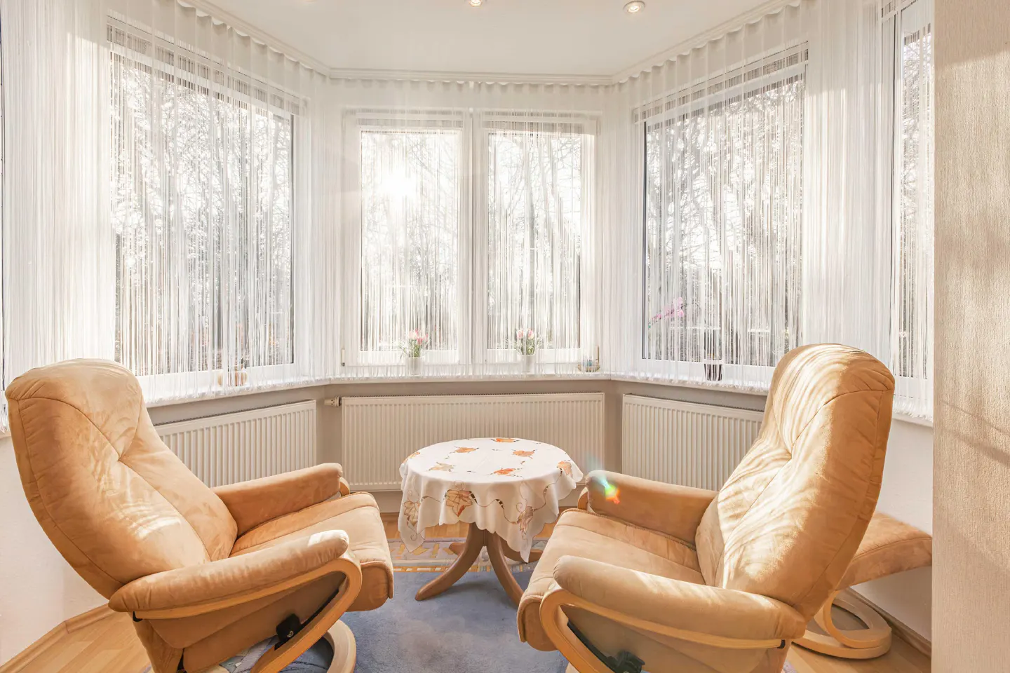 Bright sunroom with two tan recliners facing a round table with a floral tablecloth, sheer white curtains on large windows.