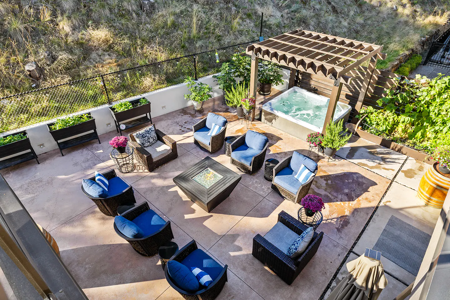 Aerial view of a patio with a hot tub, fire pit, and seating area. The patio is surrounded by greenery and a black fence.
