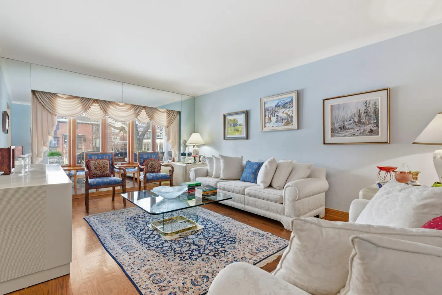 Living room with light blue walls, hardwood floors, and a blue patterned rug. White sofas and blue chairs surround a glass coffee table. Paintings hang on the wall.