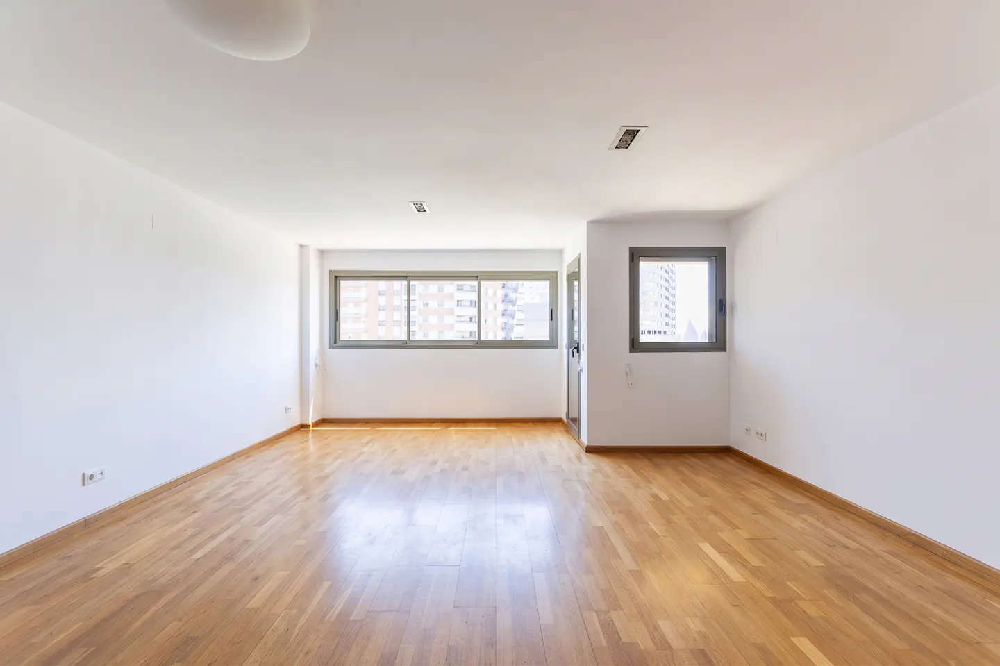 Bright, empty room with hardwood floors, white walls, and gray-framed windows showing city buildings.