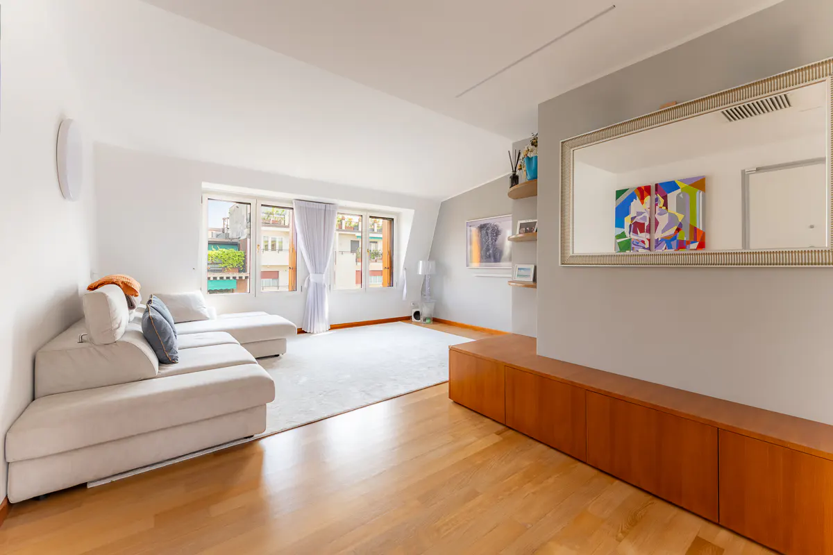 Bright living room with white walls, wood floors, and a large white sectional sofa. Windows with white curtains overlook a city street. A wooden cabinet and mirror are on the right.