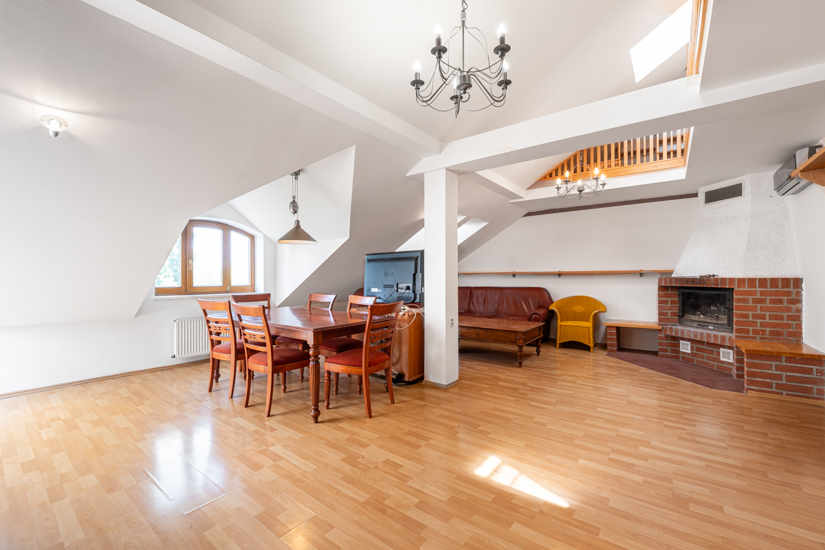Spacious attic living room with wood floors, dining set, leather sofa, brick fireplace, and vaulted ceilings with chandeliers. [AI-generated text]