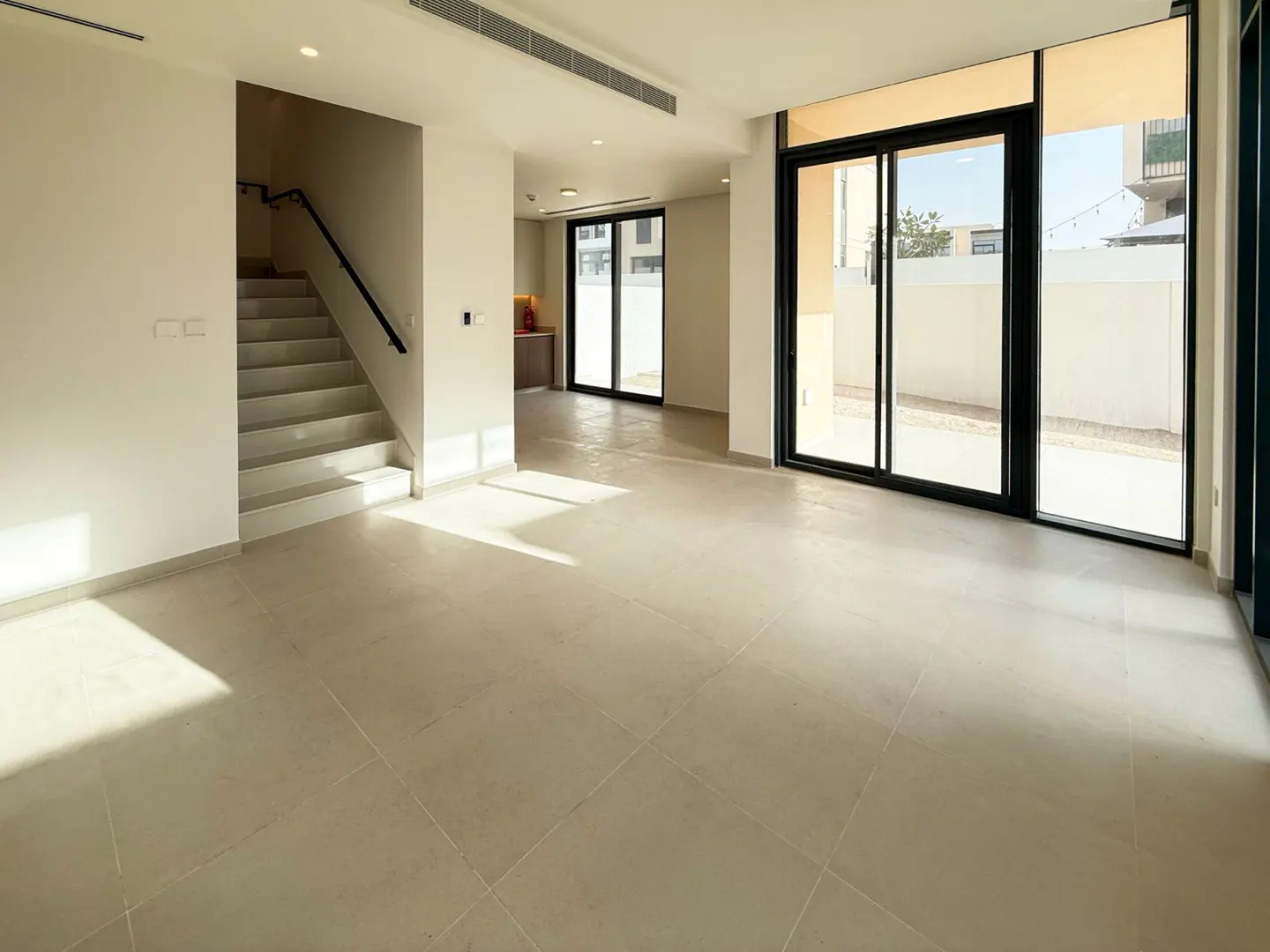 A bright, empty living room with tile floors, a staircase, and sliding glass doors to the outside.