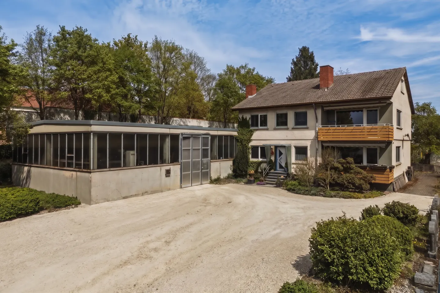 A two-story beige house with a wooden balcony next to a large, flat-roofed workshop building with a gravel driveway.