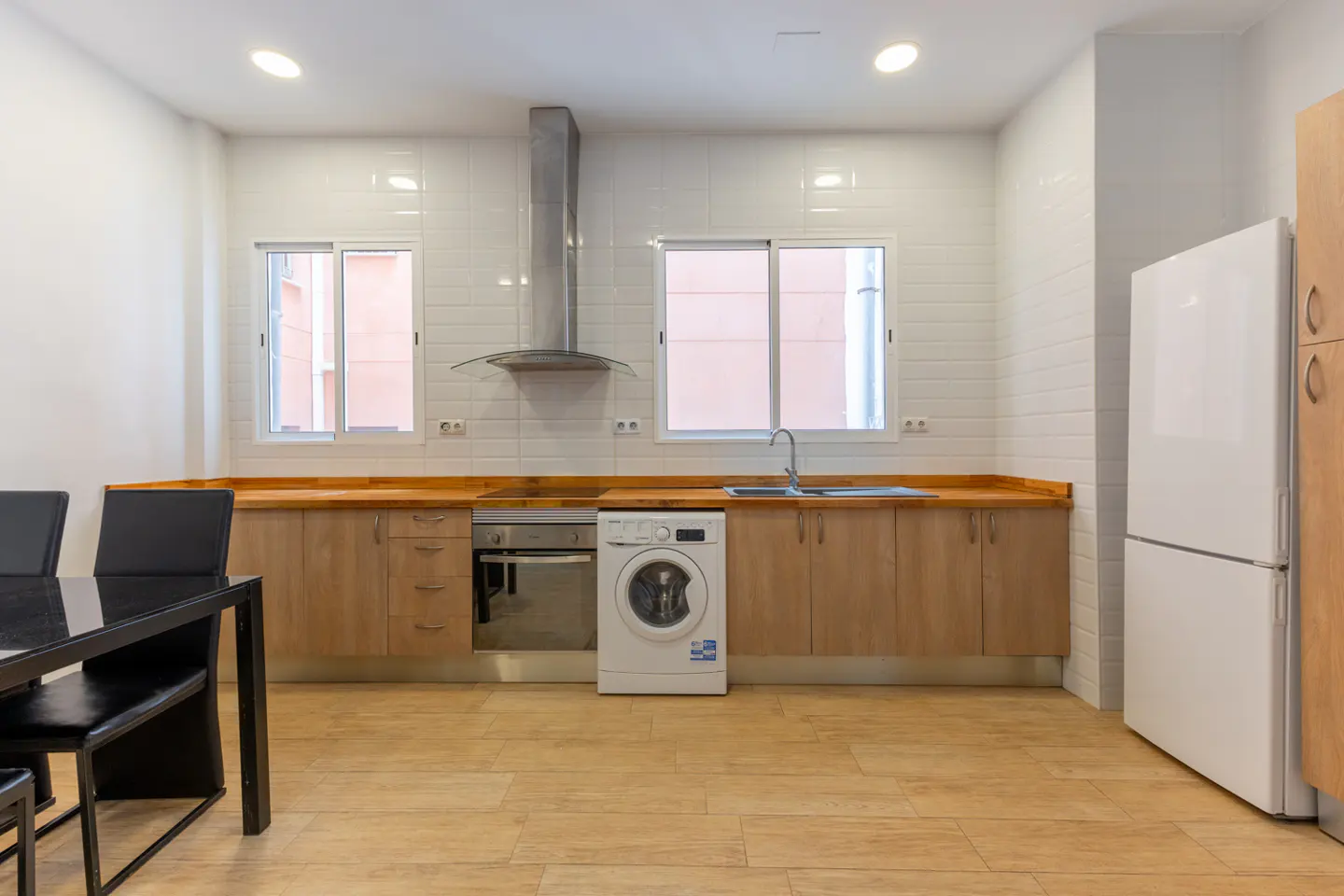 Bright kitchen with wood cabinets, stainless steel appliances, and a white washing machine. A black table and chairs are in the foreground.