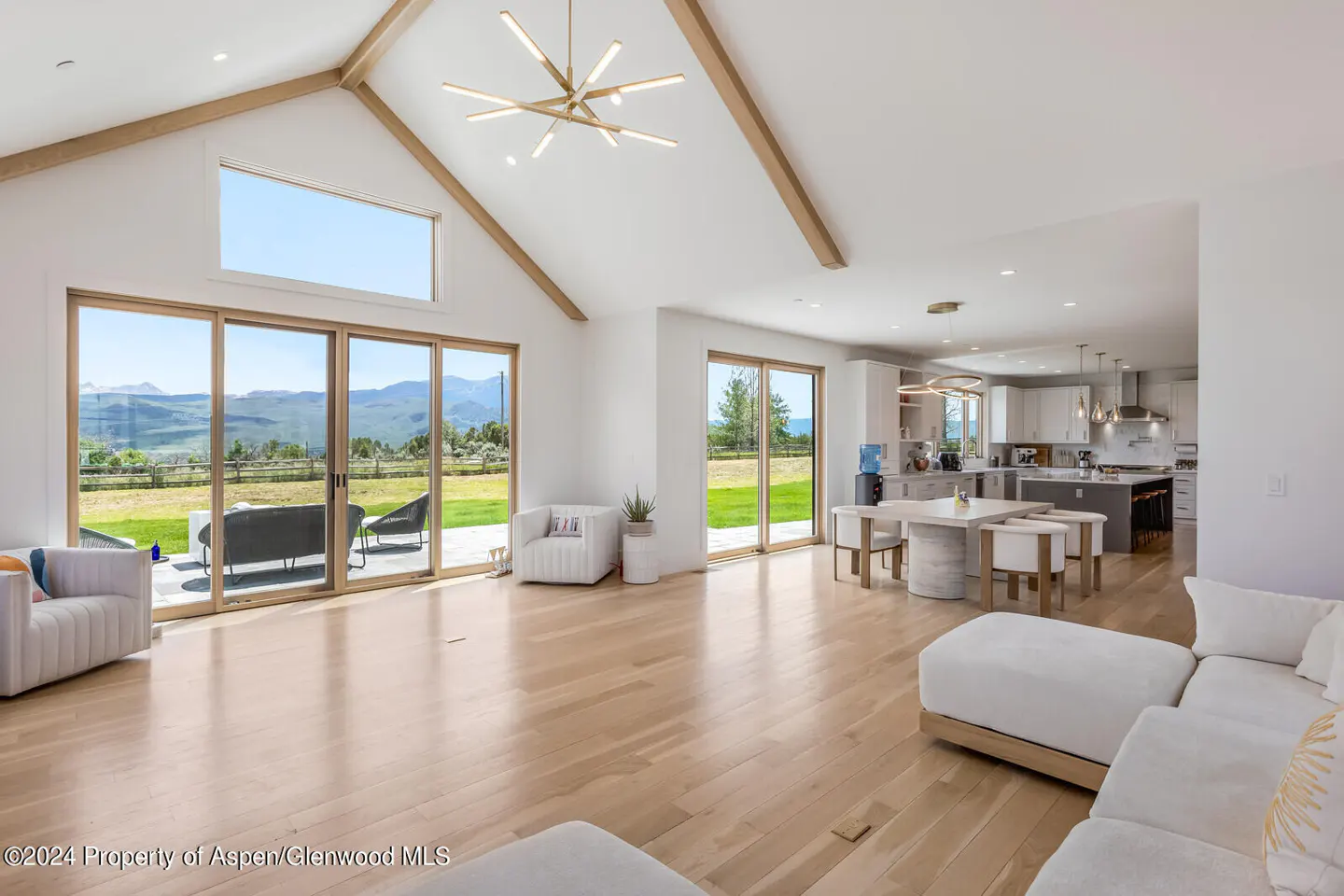 Bright, open-concept living space with white walls, wood floors, and exposed beams. Large windows offer mountain views. Modern kitchen in background.
