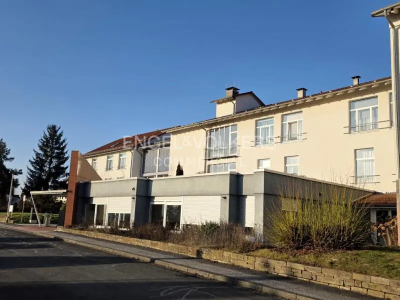 Exterior view of a multi-story beige building with white windows under a clear blue sky. Bushes and trees surround the building.