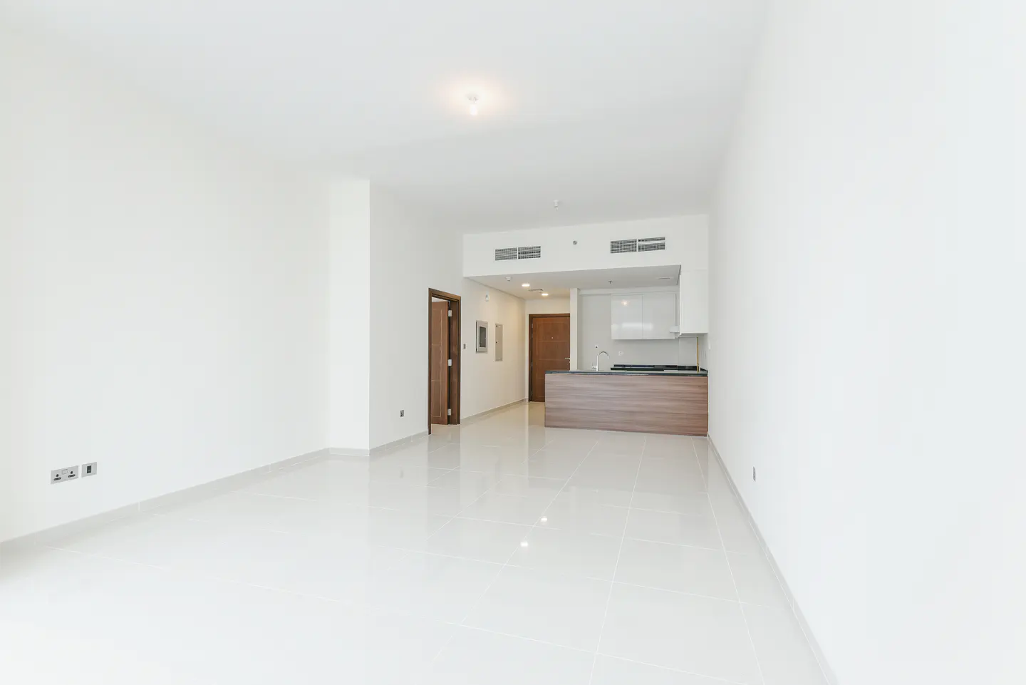 Bright, empty apartment with white walls and tile floors. A wood kitchen island and two doors are visible in the background.