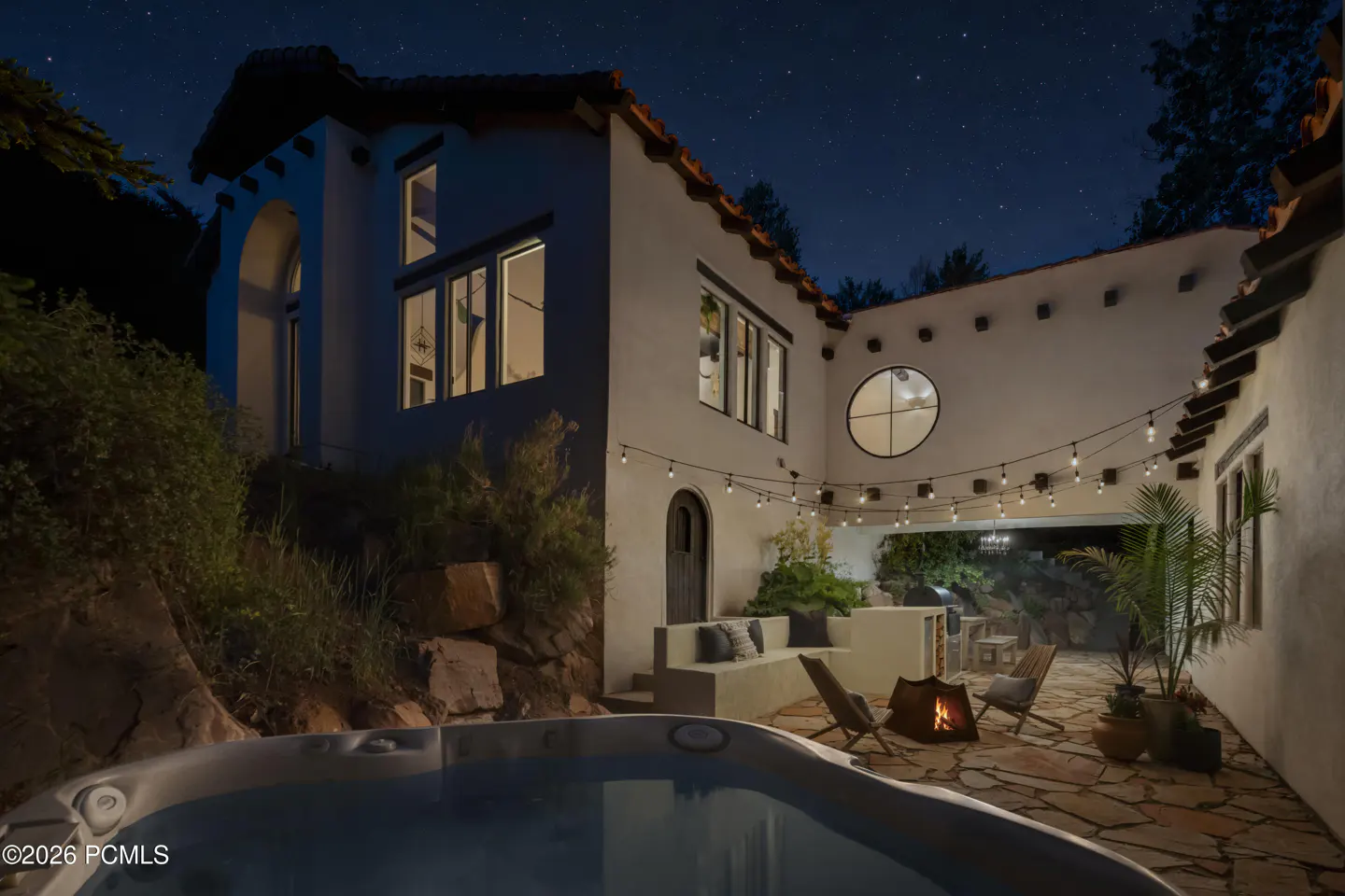 Night view of a white stucco home with a hot tub, stone patio, fire pit, and string lights under a starry sky.