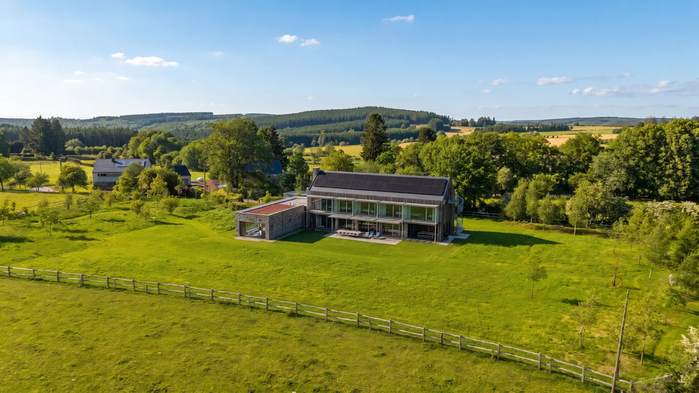 Aerial view of a modern, two-story house with a black roof and glass walls, surrounded by a green lawn, trees, and a wooden fence.