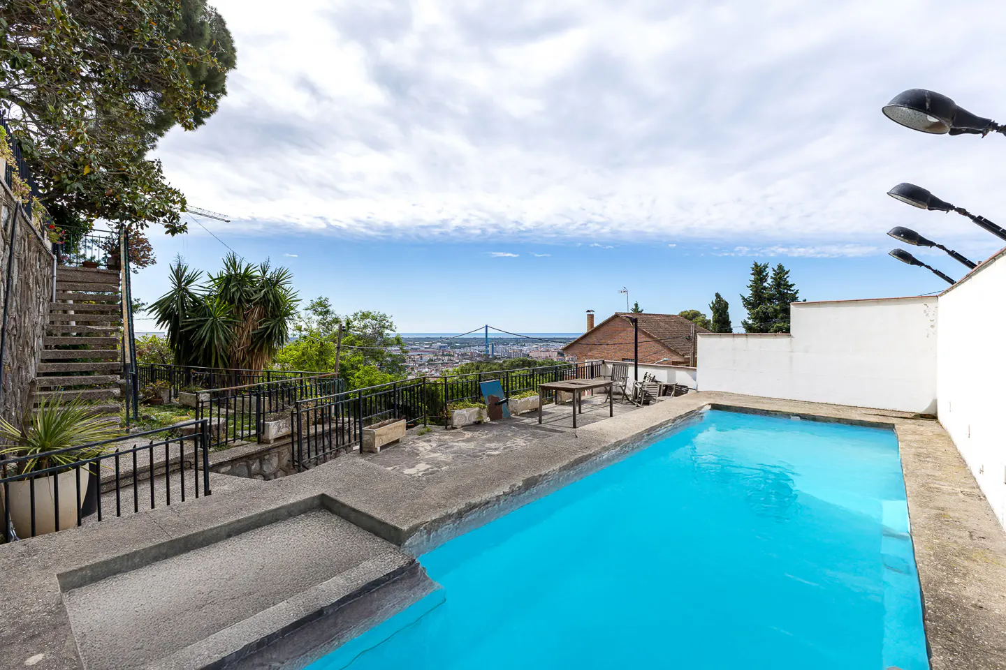 Outdoor pool with turquoise water, concrete surround, and city view with a bridge in the distance under a cloudy sky.