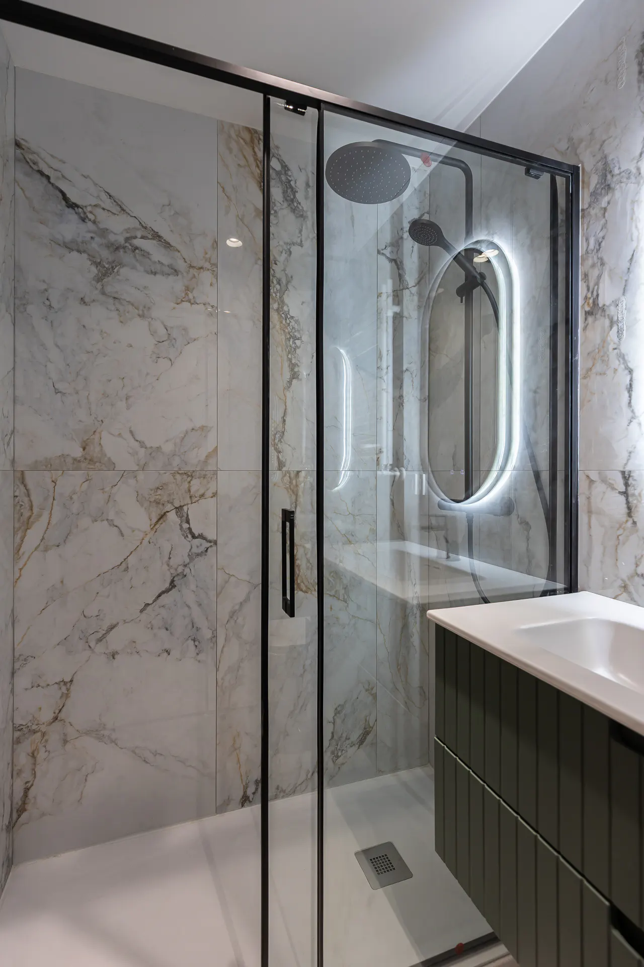 Modern bathroom with marble walls, black framed glass shower, rain shower head, and an oval mirror with LED lighting. A green vanity with a white countertop is visible.